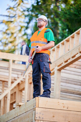 Carpenter building wooden frame two-story house. Bearded man in glasses holding a level, wearing protective helmet, overalls and orange vest. Concept of modern ecological construction.