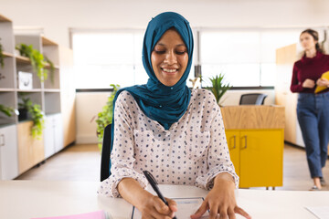 Happy biracial casual businesswoman with hijab writing and having video call in creative office