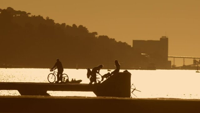 Silhouette of people have rest in park or pier near river bridge - man on bycicle and family park near river in Almada, Portugal - sunset
