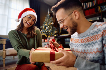 Happy woman surprising her boyfriend with Christmas present at home. - obrazy, fototapety, plakaty