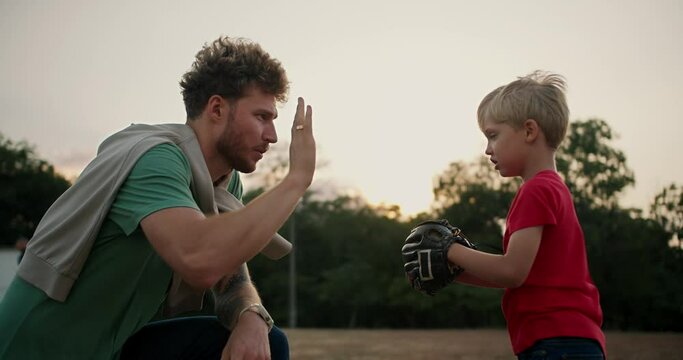 Dad with curly hair and stubble in a Green T-shirt helps his little son Blondie in a red T-shirt put on a baseball glove and high-fives Him in the park
