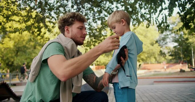 A man with curly hair and stubble in a green T-shirt helps his son in a blue sweater put on his elbow pads before riding a skateboard at a skatepark in the park. Little blond boy preparing to ride a