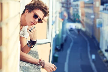 Poster Portrait, fashion or apartment window with a man looking out at a city street in sunglasses in summer. Face, space or urban style and a confident or cool young model in trendy shades and accessories © Nicola Katie/peopleimages.com