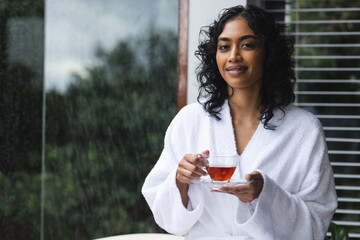 Happy biracial woman in bathrobe sitting on bath with tea and looking on camera in sunny bathroom