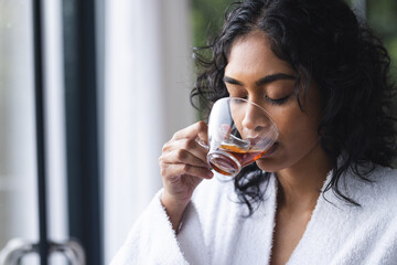 Happy biracial woman in bathrobe drinking tea in sunny room at home