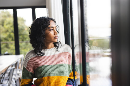 Worried biracial woman standing by window and looking ahead in sunny living room