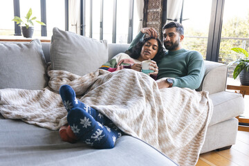Biracial couple embracing on sofa under blanket and drinking cocoa in living room at home
