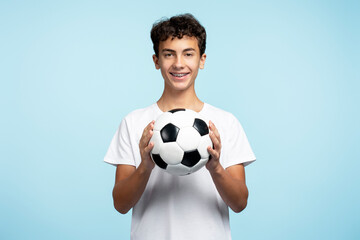 Portrait of smiling teenage boy holding ball playing football isolated on blue background. Sport,...