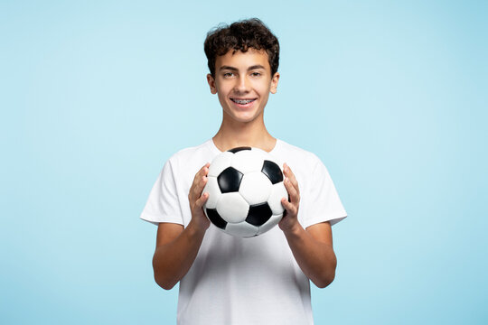 Portrait of smiling teenage boy holding ball playing football isolated on blue background. Sport, hobby, competition concept