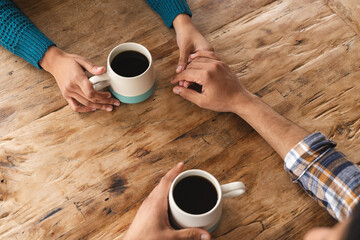 Hands of biracial couple sitting at table with coffee in living room at home