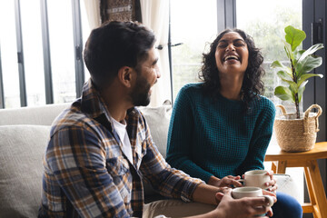 Happy biracial couple sitting on sofa with coffee and smiling in living room at home