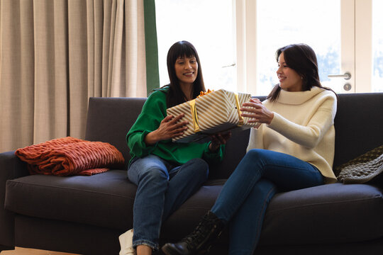 Happy diverse mother and daughter sitting on couch, sharing presents at home, copy space