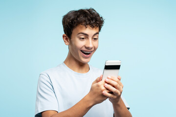 Portrait of overjoyed teenage boy with braces holding smartphone playing mobile game, communication...