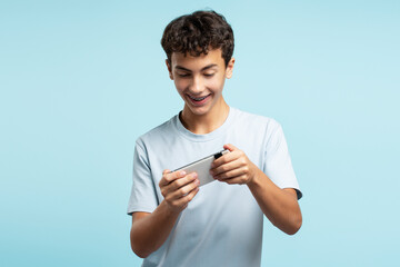 Young happy boy holding mobile phone playing mobile game isolated on blue background 