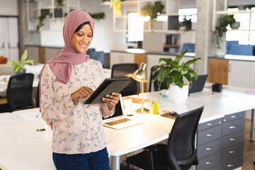Happy biracial casual businesswoman with hijab using tablet in creative office, copy space