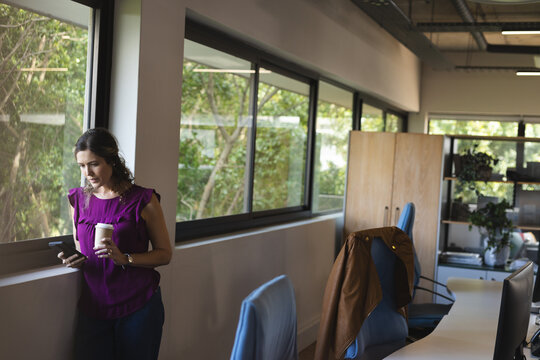 Happy caucasian casual businesswoman with takeaway coffee talking on smartphone in office
