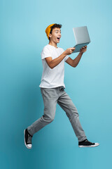 Overjoyed boy teenager holding laptop computer playing video game,  jumping isolated on blue...