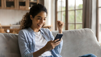 Happy excited young latin woman relaxing on couch using phone winning money in online app  - obrazy, fototapety, plakaty