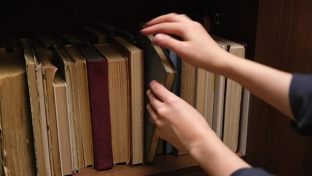 A bookshelf with old books in the closet. A woman hand goes through the spines of books in search of a suitable book, pulling, replacing. Slow motion