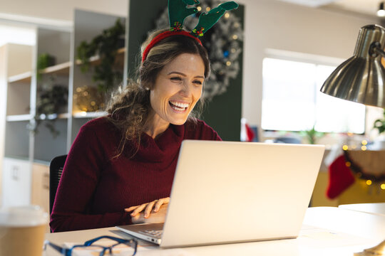 Happy caucasian casual businesswoman wearing reindeer antlers having laptop video call in office