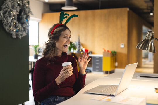 Happy caucasian casual businesswoman wearing reindeer antlers having laptop video call in office