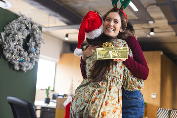 Happy diverse female colleagues wearing christmas hats giving presents and embracing in office