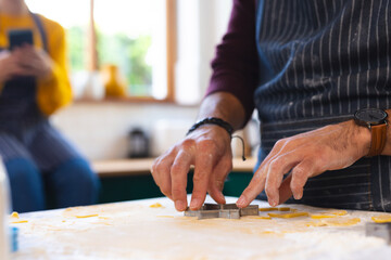 Midsection of caucasian couple cutting cookie dough and using smartphone in kitchen, copy space