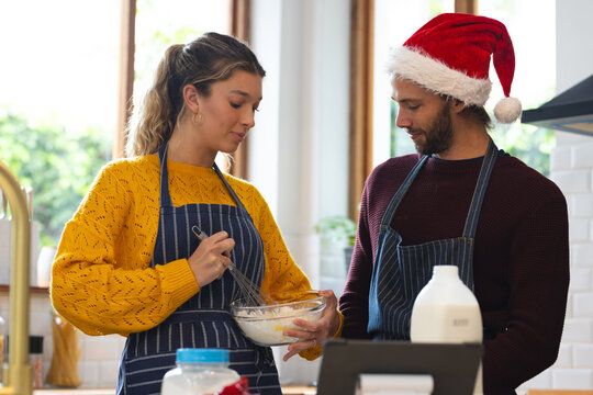Happy caucasian couple in aprons and christmas hat baking and using tablet in kitchen