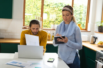 Focused caucasian couple discussing bills, making notes and using laptop in kitchen