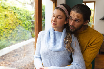 Happy, romantic caucasian couple embracing and smiling by window at home