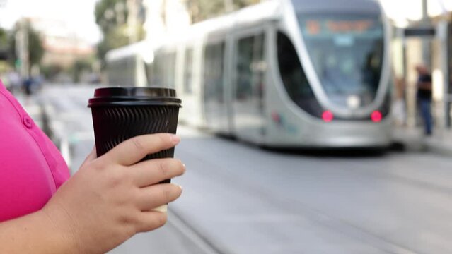 A close-up of a woman in a pink shirt holding a black plastic coffee cup in her hand with a passing tram and a bustling cityscape in the background on a sunny summer day