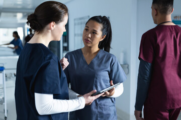 Diverse female doctors discussing work, using tablet in corridor at hospital