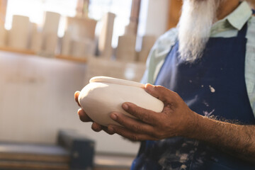 Mid section of senior biracial potter with beard, holding clay vase in pottery studio
