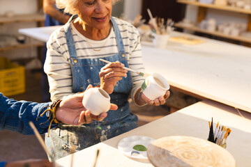 Happy biracial senior female potter with gray hair, glazing clay jug and smiling in pottery studio