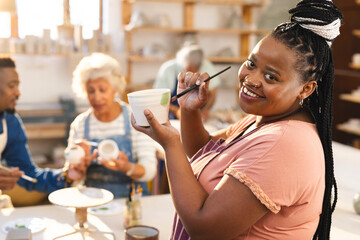 Happy african american female potter glazing clay jug with others in pottery studio