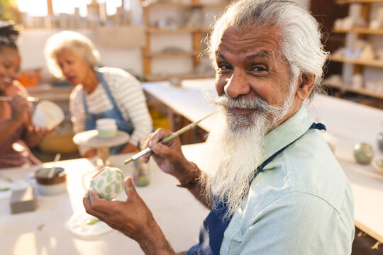 Happy biracial senior potter with long beard glazing clay jug and smiling in pottery studio