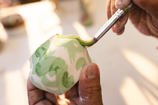 Hands of biracial senior potter glazing clay jug in pottery studio