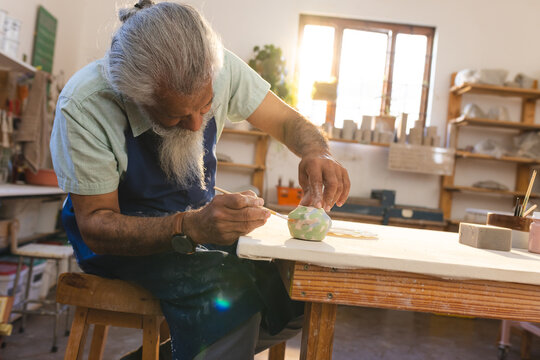 Focused biracial senior potter with long beard glazing clay jug in pottery studio