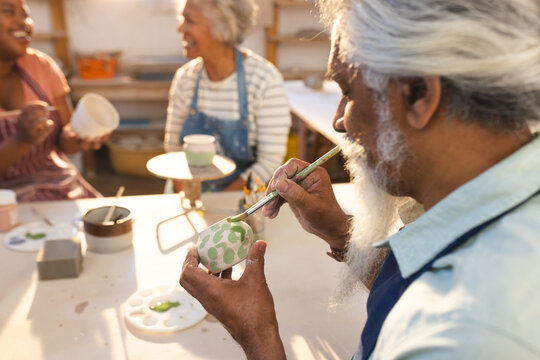 Focused biracial senior potter with long beard glazing clay jug in pottery studio