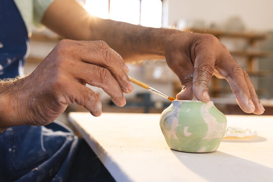 Hands of biracial senior male potter with glazing clay jug in pottery studio