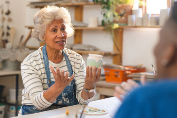 Happy biracial senior female potter with gray hair, discussing with others in pottery studio