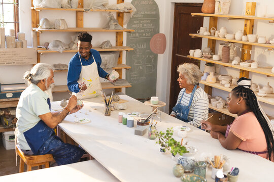 Happy diverse group of potters glazing clay jugs and discussing in pottery studio
