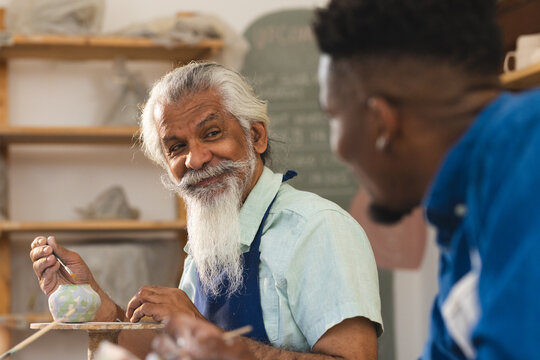 Two diverse male potters glazing clay jug and discussing in pottery studio