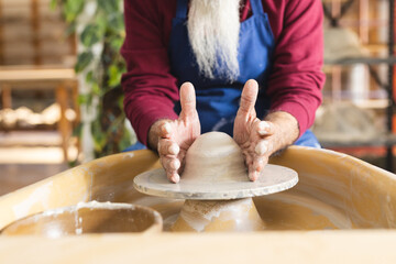 Focused biracial senior potter with long beard using potter's wheel in pottery studio