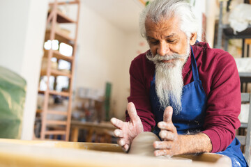Focused biracial senior potter with long beard using potter's wheel in pottery studio