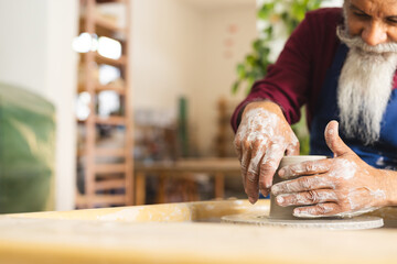 Focused biracial senior potter with long beard using potter's wheel in pottery studio