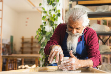 Focused biracial senior potter with long beard using potter's wheel in pottery studio