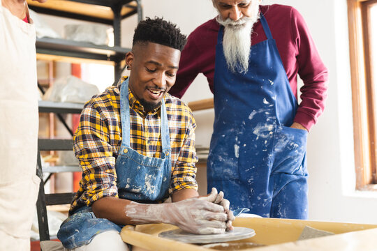 Happy african american potter using potter's wheel with others in pottery studio