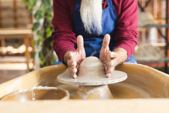 Focused biracial senior potter with long beard using potter's wheel in pottery studio