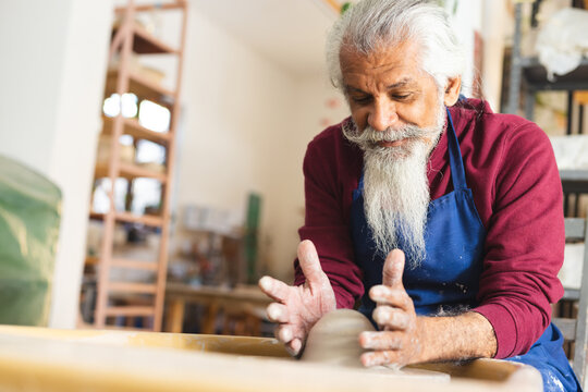 Focused biracial senior potter with long beard using potter's wheel in pottery studio